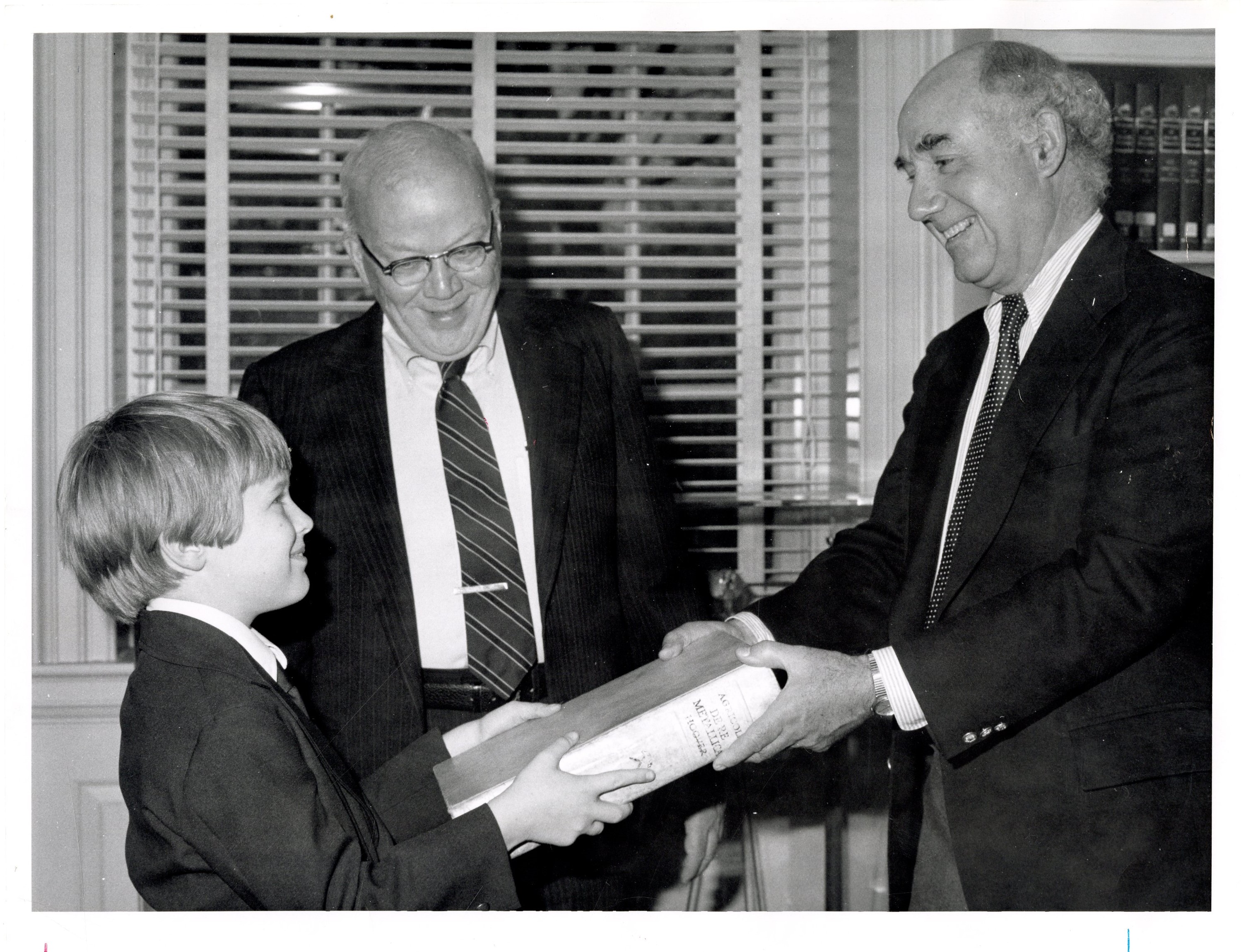 black and white photo of two men standing exchanging book with small boy