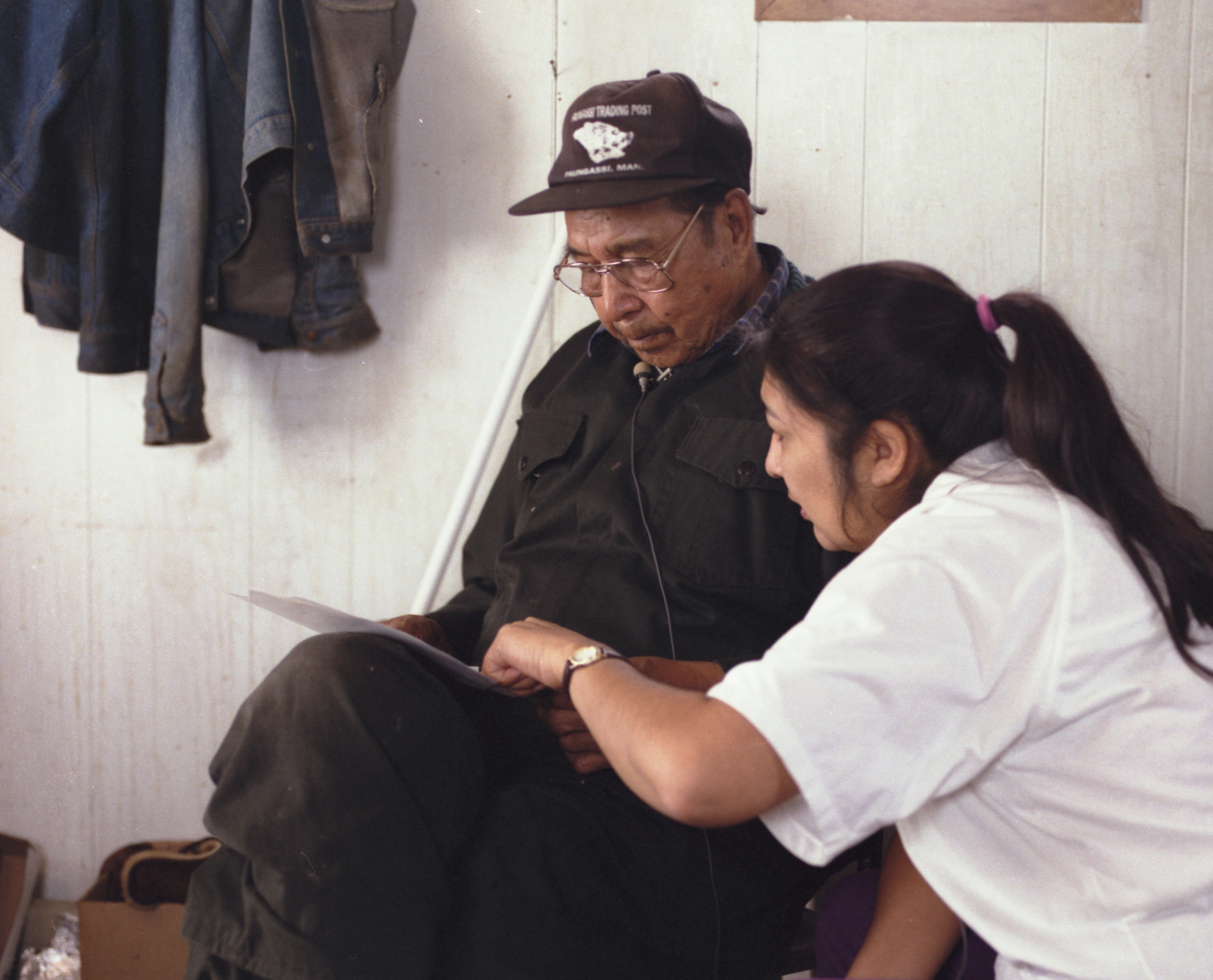 Fotografía en color de un hombre sentado con una gorra de béisbol y ropa oscuras, y una mujer con una cola de caballo y una camisa blanca, juntos mirando hojas de papel.