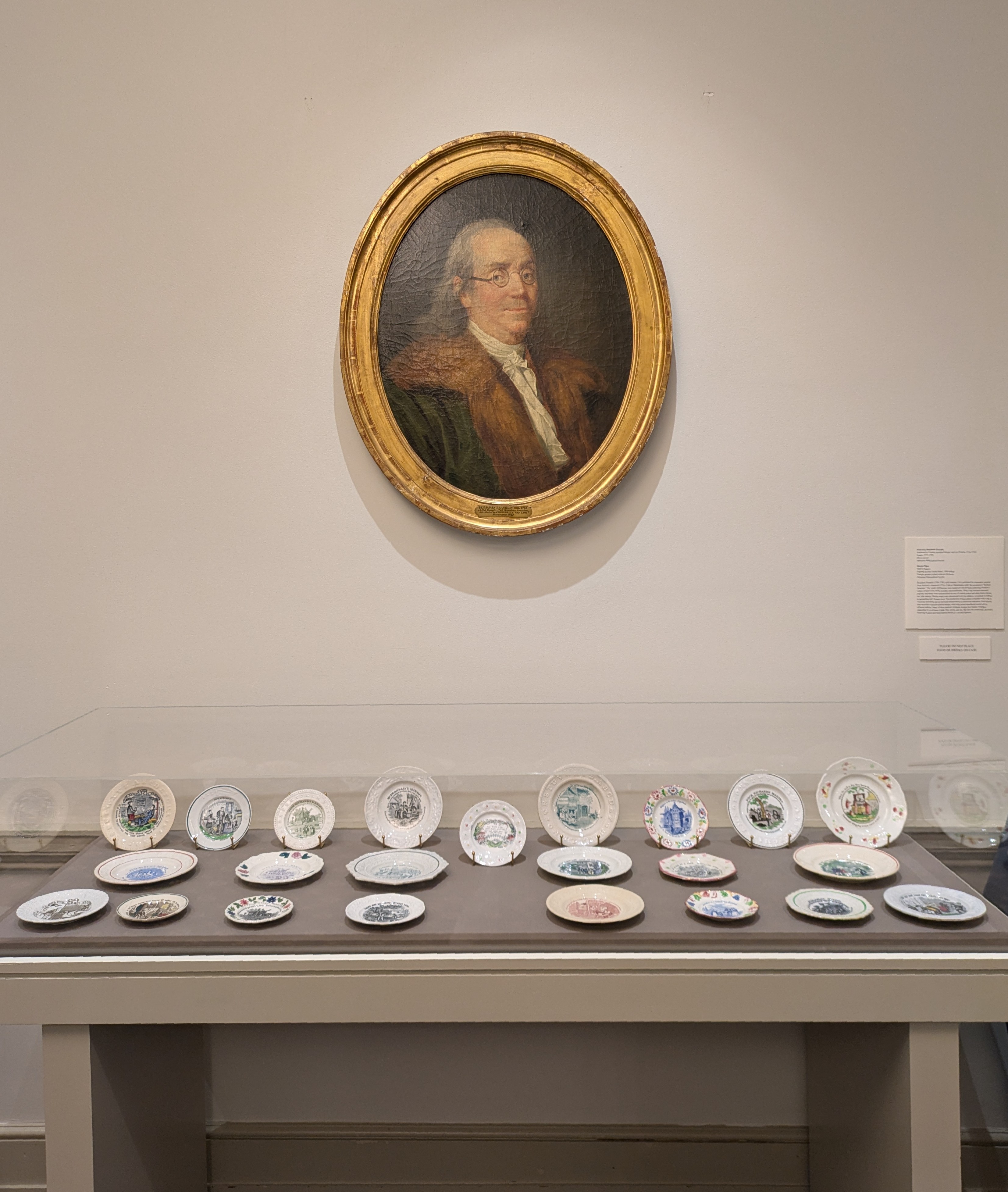 photo of decorative plates arranged in display case with portrait of Benjamin Franklin hanging over