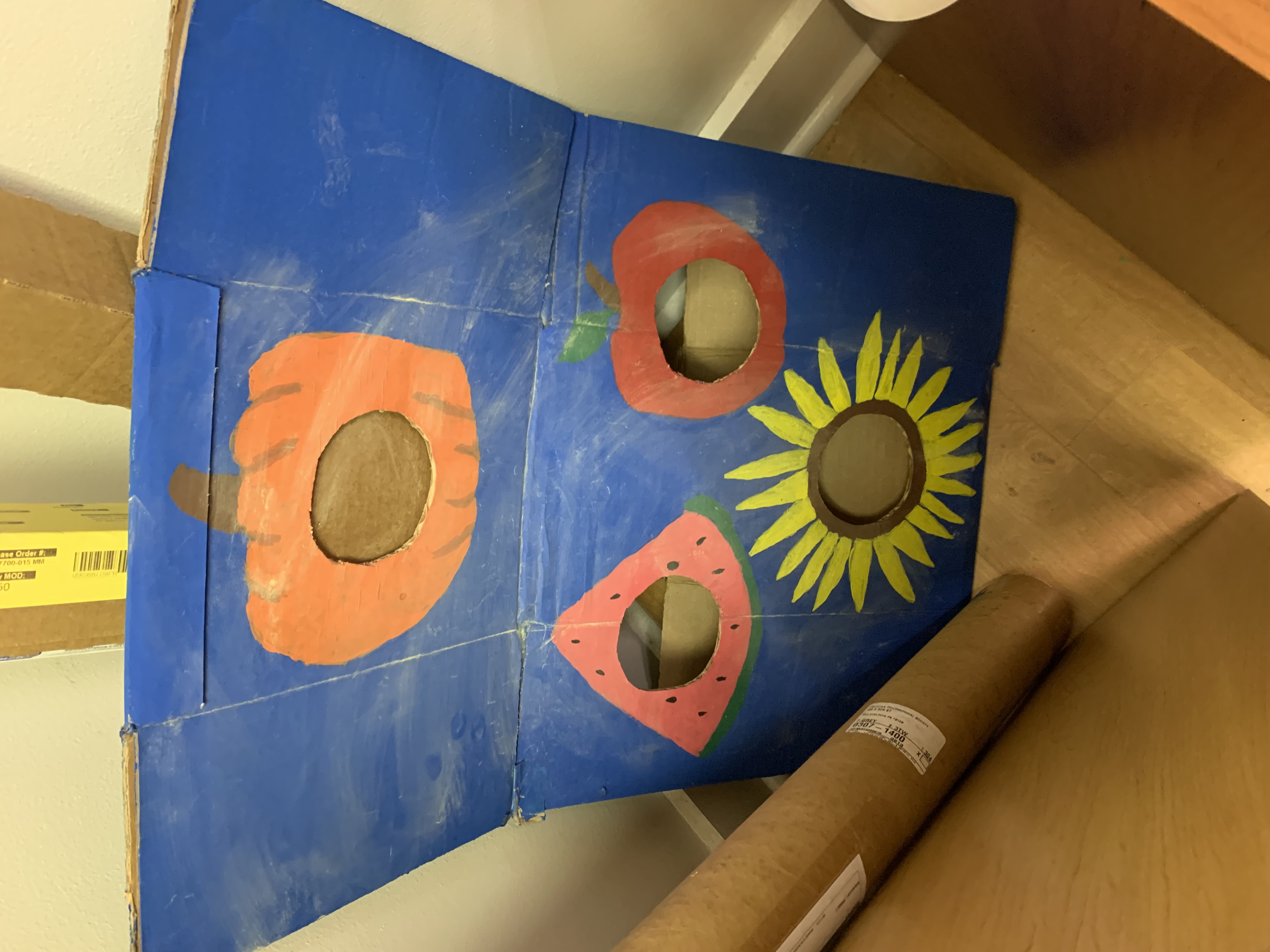 photo of pollination cornhole game. painted cardboard (blue background) with pumpkin, watermelon, apple, and sunflower "holes" painted on