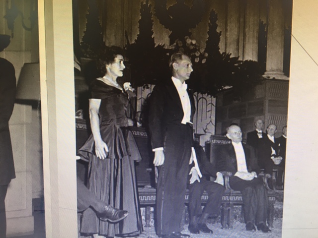 Black and white photo of Gerty (left) and Carl (right) Cori standing on a stage with others seated behind them