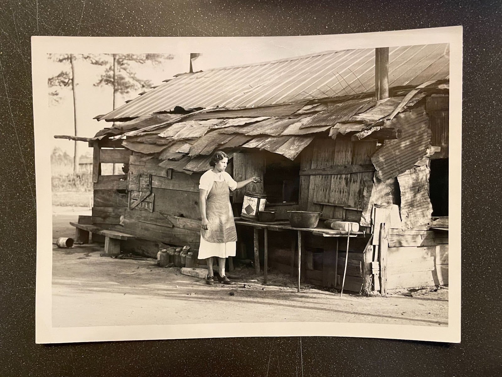 Fotografía de una mujer vestida con un vestido y un delantal, parada frente a una vivienda rústica.