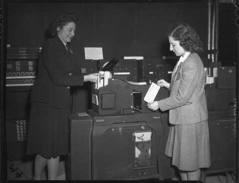 black and white photograph of two women standing, operating the punch card machine