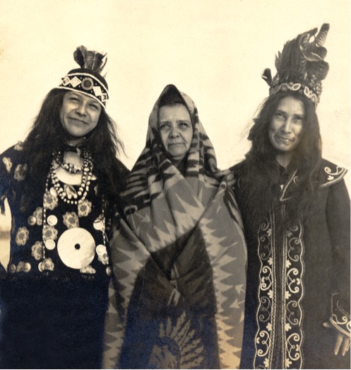 Fotografía sepia de tres mujeres paradas juntas mirando a la cámara, vestidas con atuendos ornamentados.
