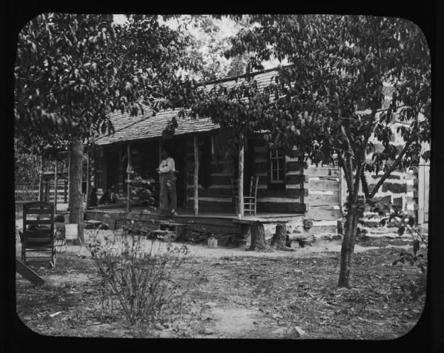 black and white photo of man on porch of log cabin