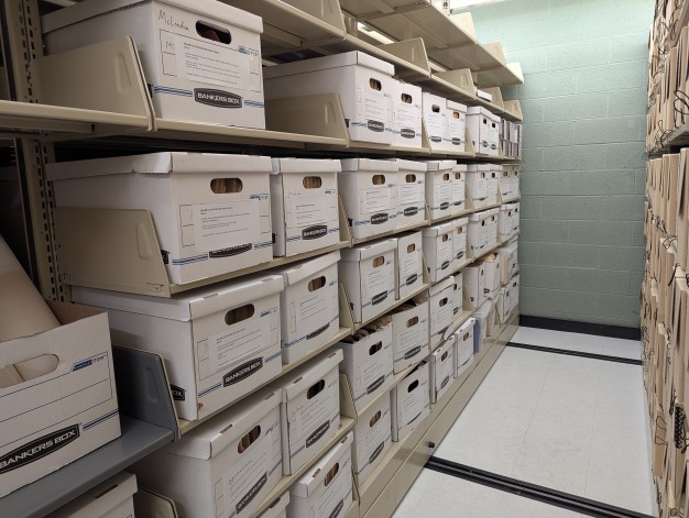 A photo of a corridor flanked by two library rolling shelving units. The unit on the left is filled with white banker’s boxes with typed labels taped to each box.