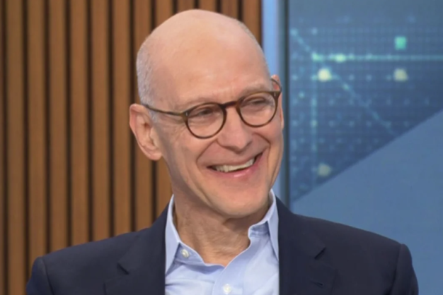 A man with black-framed glasses smiles on a news set.