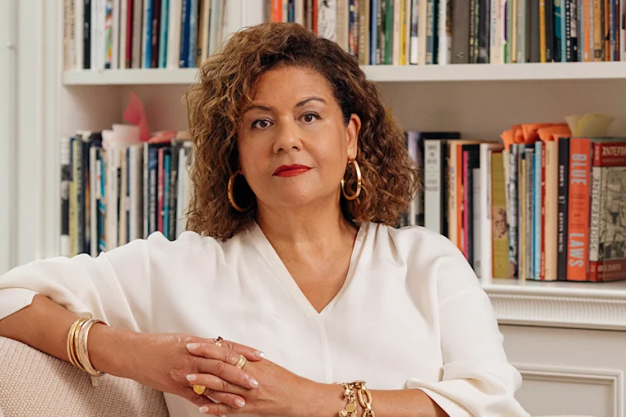 A woman in a white shirt sits in front of a bookshelf.