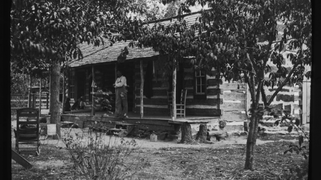 black and white photo of man on porch of log cabin