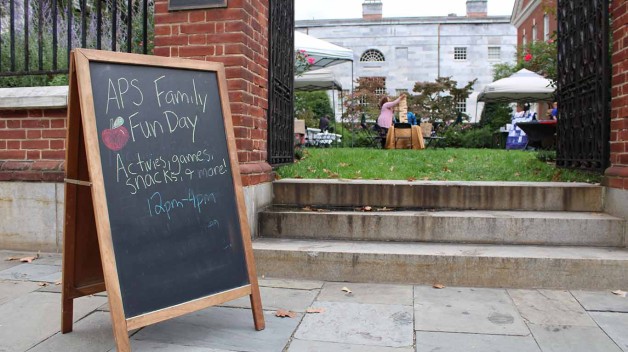 Image of a chalkboard with the words "APS Family Fun Day activities, games, snacks, & more! 12pm - 4pm." There is also a drawing of a red apple.  Behind the sign is the entrance to Jefferson Garden and people participating in activities. 