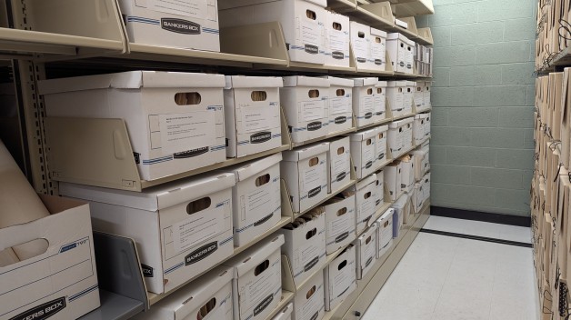 A photo of a corridor flanked by two library rolling shelving units. The unit on the left is filled with white banker’s boxes with typed labels taped to each box.