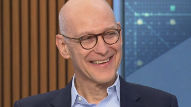 A man with black-framed glasses smiles on a news set.