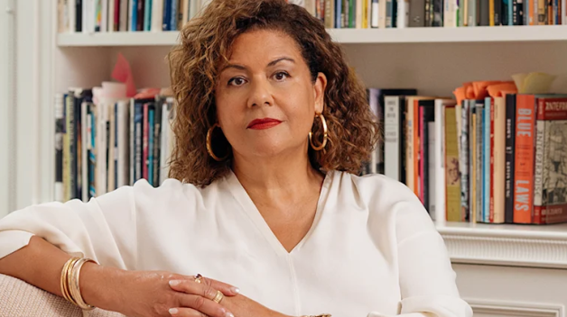 A woman in a white shirt sits in front of a bookshelf.
