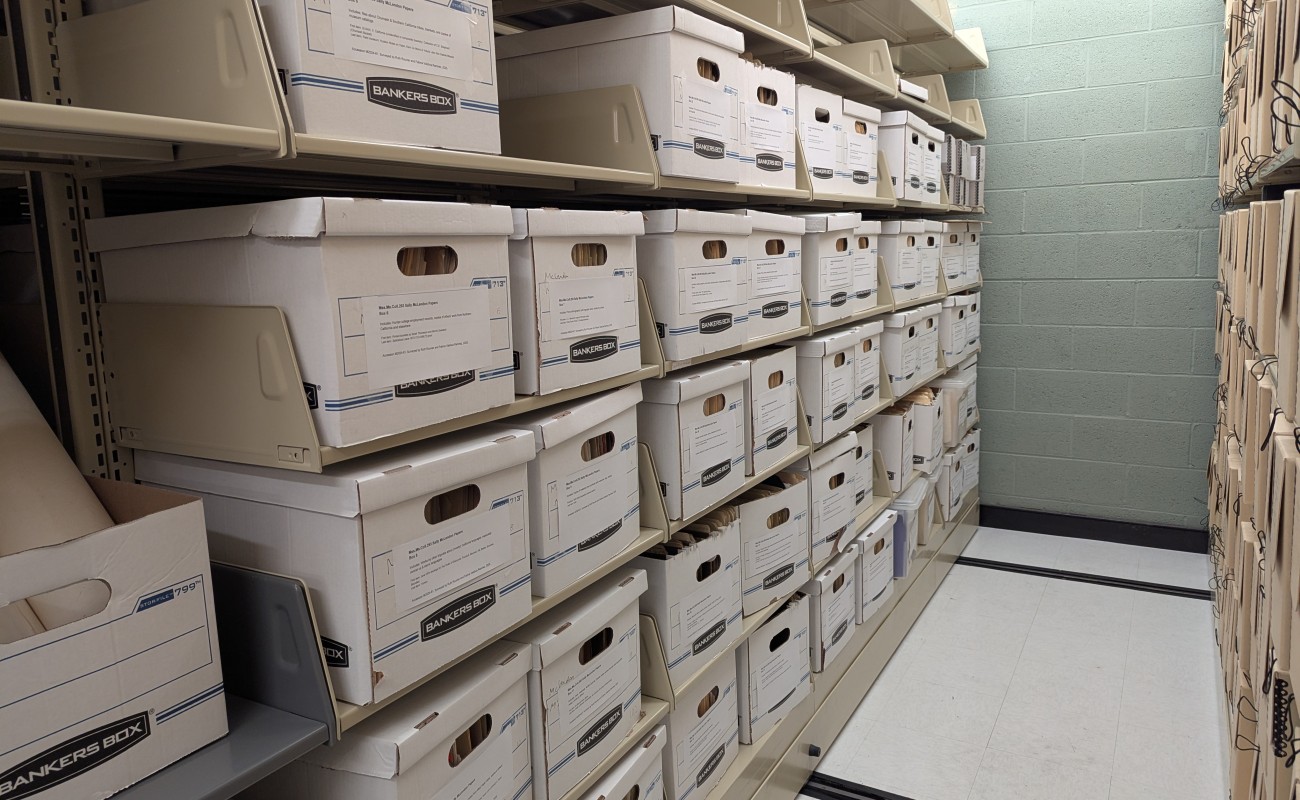 A photo of a corridor flanked by two library rolling shelving units. The unit on the left is filled with white banker’s boxes with typed labels taped to each box.