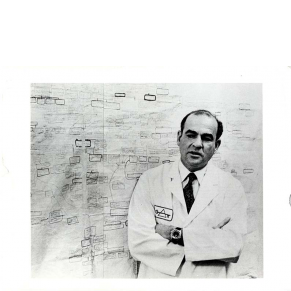 black and white photograph of a man (Blumberg) wearing a lab coat standing in front of a chart