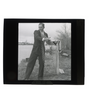 Black-and-white glass lantern slide of a Houma man wearing a suit and spreading out a fishing net.