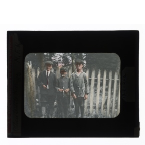 Hand-colored glass lantern slide of three Huron-Wendat boys, in the attire of the day, smoking by a fence, Ontario, Canada.
