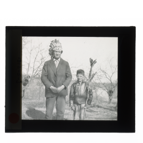 Black-and-white glass lantern slide man wearing head dress standing next to boy with spear.