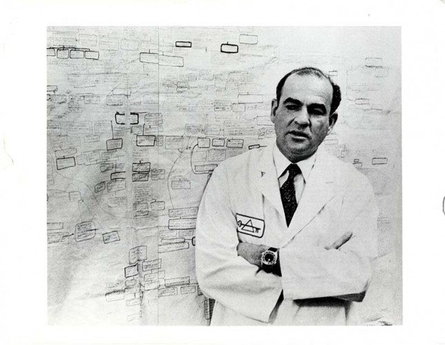 black and white photograph of a man (Blumberg) wearing a lab coat standing in front of a chart