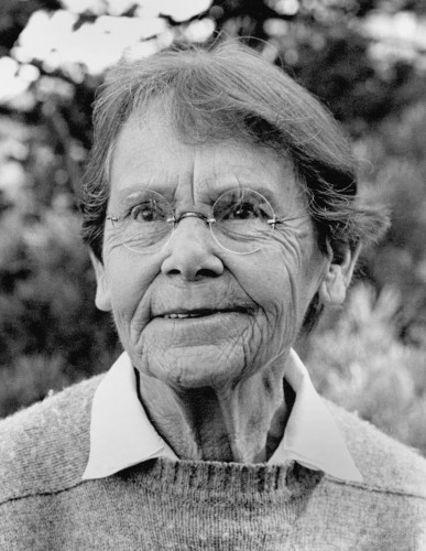 black and white photograph of a woman with short hair wearing a collared shirt and glasses standing in front of trees