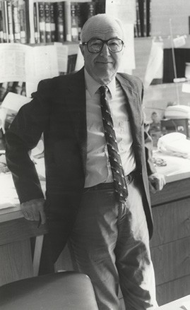 black and white photograph of a man in a suit and tie facing the camera and leaning against a desk