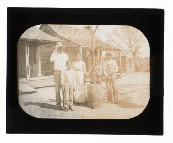 Black-and-white glass lantern slide of Creek family, girl posing with mortar and pestle, boy posing with bow and arrow.