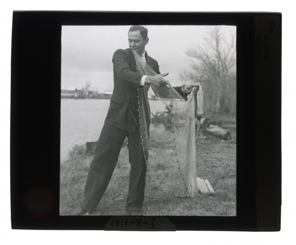 Black-and-white glass lantern slide of a Houma man wearing a suit and spreading out a fishing net.