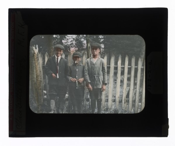 Hand-colored glass lantern slide of three Huron boys, in the attire of the day, smoking by a fence, Ontario, Canada.