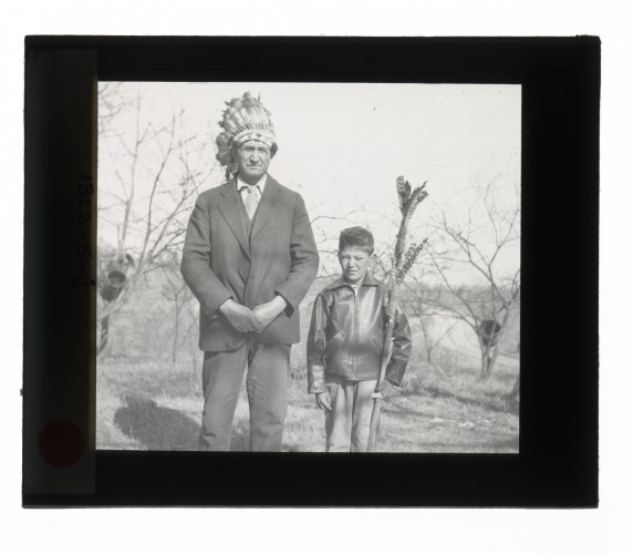 Black-and-white glass lantern slide man wearing head dress standing next to boy with spear.