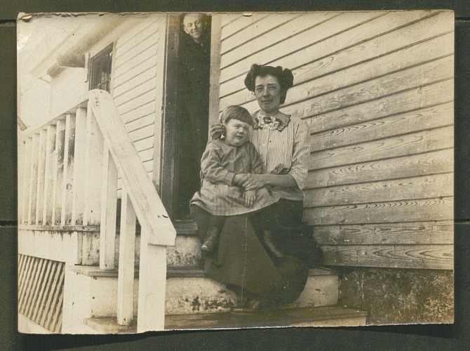 black and white photo of women and child seated on stairs with man peeking out of door in background